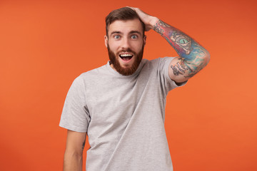 Amazed bearded brunette man with tattooes and short haircut looking at camera with wide mouth opened and rounding his blue eyes, holding raised hand on his head while standing over orange background