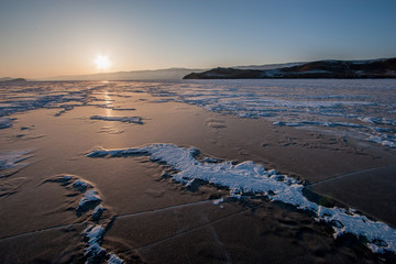 Ice of Lake Baikal at sunset with cracks and snow. Mountains beyond the lake. The sun is on the horizon. Reflection of the sun on ice.