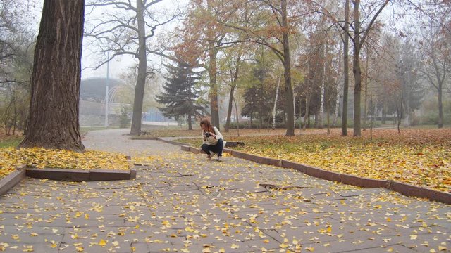 Alone young girl in white coat and backpack taking pictures in a foggy city park. yellow autumn leaves lie around the girl on the ground