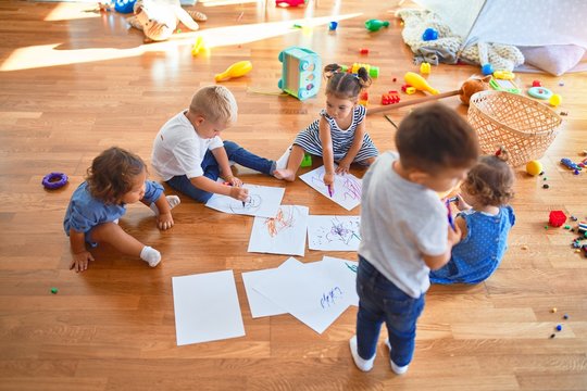 Adorable group of toddlers sitting on the floor drawing using paper and pencil around lots of toys at kindergarten
