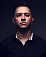 Obraz premium Sexy young man looking serious in black shirt on grey studio dark shadow background. Closeup toned color portrait