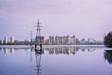Fototapeta na wymiar High-voltage power line over the wide lake and city in the distance. Sunset and reflection in water. Electrical conductor. Technology in the city. Voronezh, Russia.