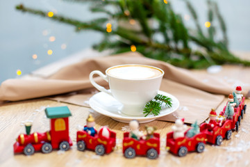 Delicious fresh festive morning cappuccino coffee in a ceramic white cup on the wooden table with decorative christmas train, red ornamentals, fireflies and spruce branches