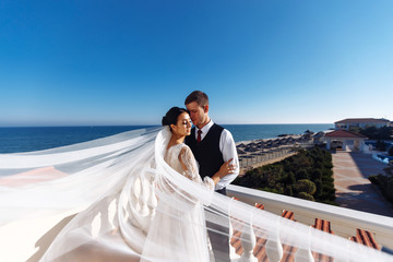 Newlyweds enjoy each other tenderly in the shadow of a flying veil on the background of a beautiful sea landscape. The bride and groom hugging and kissing on the background of the sea. 