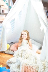 Adorable blonde toddler playing sitting on blanket inside tipi at kindergarten
