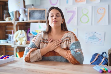 Young beautiful teacher woman wearing sweater and glasses sitting on desk at kindergarten smiling...