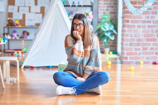 Young beautiful teacher woman wearing sweater and glasses sitting on the floor at kindergarten looking stressed and nervous with hands on mouth biting nails. Anxiety problem.