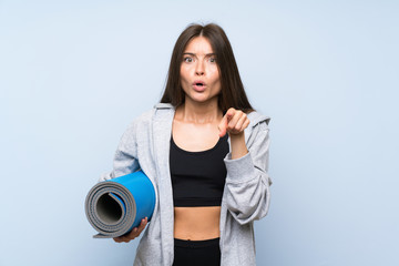 Young sport girl with mat over isolated blue background surprised and pointing front