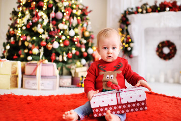 a child-a little boy in a red Christmas sweater with a picture of Santa's reindeer sits barefoot on a warm red carpet with a gift in his hands on the background of a Christmas tree and decor