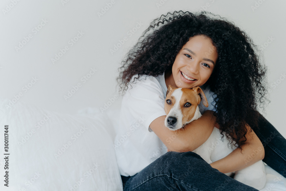 Wall mural Photo of attractive young woman with Afro hircut, embraces with love dog, takes care of pet, smiles gently, wears casual clothing, isolated over white background, sit on bed, copy space for your promo