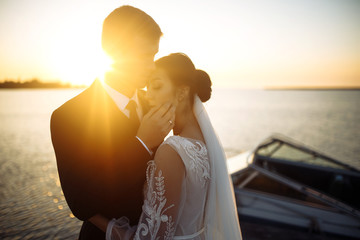 Beautiful bride and stylish groom together on the bridge against the background of the boat at sunset. Newlyweds tenderly hug, kiss and enjoy each other at sunset. Wedding. Love. Romantic moment. 