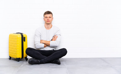 Young handsome man sitting on the floor with a suitcase keeping arms crossed