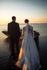 Beautiful bride and stylish groom together on the bridge against the background of the boat at sunset. Newlyweds tenderly hug, kiss and enjoy each other at sunset. Wedding. Love. Romantic moment. 