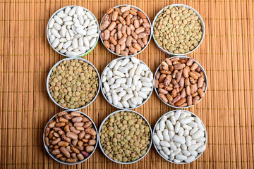 Nine round bowls with organic fresh green lentil beans, red and white beans on textured bamboo wooden background table in warm light, top view or flat lay of healthy food