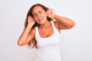 Middle age mature woman standing over white isolated background Trying to hear both hands on ear gesture, curious for gossip. Hearing problem, deaf