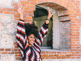 A pretty afro woman raises her arms and smiles, while leaning on a brick wall next to a window