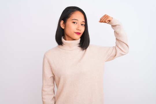 Young chinese woman wearing turtleneck sweater standing over isolated white background Strong person showing arm muscle, confident and proud of power