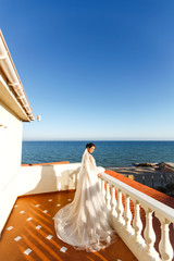 Beautiful bride with dark hair in luxurious wedding dress posing in balcony with sea view. Happy bride with luxury make-up and hairstyle. Luxurious elegant bride in a white dress in the sun.