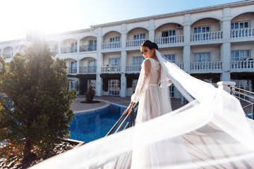 Obraz premium Beautiful sexy bride in white dress posing near the pool. Happy bride in luxurious wedding dress posing in the sunshine. Wind blows her veil. Wedding day. Marriage. Fashion bride.
