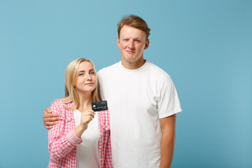 Young smiling couple two friends guy girl in white pink empty blank t-shirts posing isolated on pastel blue background in studio. People lifestyle concept. Mock up copy space. Hold credit bank card.