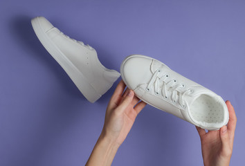 Female hands hold white stylish sneakers on purple studio background. Top view.