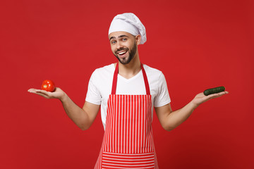 Young bearded male chef cook or baker man in striped apron white t-shirt toque chefs hat isolated on red background. Cooking food concept. Mock up copy space. Holding fresh vegetables tomato cucumber.