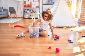 Beautiful toddler wearing glasses and unicorn diadem sitting on the floor playing with building blocks at kindergarten