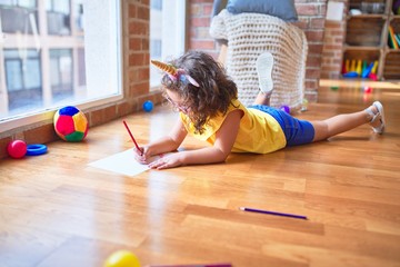 Beautiful toddler wearing glasses and unicorn diadem lying down on the floor drawing using paper...