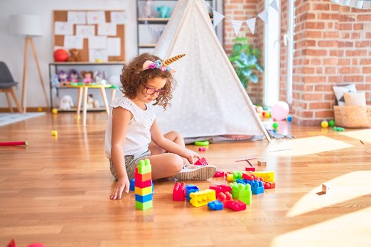 Beautiful toddler wearing glasses and unicorn diadem sitting playing with building blocks at kindergarten