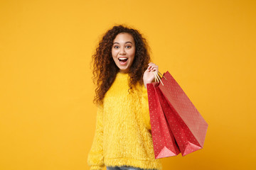 Surprised young african american girl in fur sweater posing isolated on yellow orange wall background. People lifestyle concept. Mock up copy space. Holding package bag with purchases after shopping.