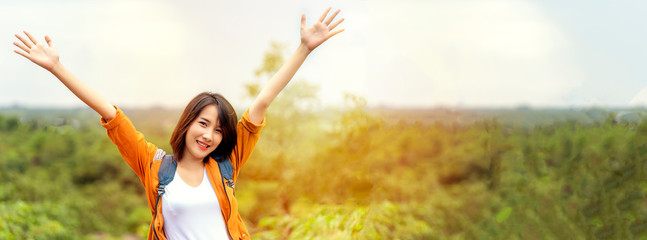 Young attractive woman raising hands up in air with smiling face. Feeling freedom, motivation with energy while climbing to top of mountain with forest landscape background, leisure backpacker