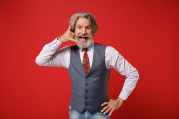 Cheerful elderly gray-haired mustache bearded man in classic shirt vest tie posing isolated on red background. People lifestyle concept. Mock up copy space. Doing phone gesture like says call me back.