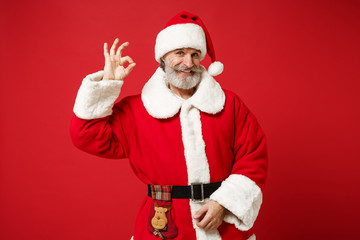 Fototapeta na wymiar Smiling elderly gray-haired mustache bearded Santa man in Christmas hat posing isolated on red wall background. Happy New Year 2020 celebration holiday concept. Mock up copy space. Showing Ok gesture.