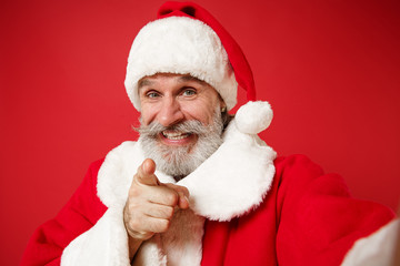 Close up of elderly gray-haired mustache bearded Santa man in Christmas hat posing isolated on red background. New Year 2020 celebration concept. Mock up copy space. Doing selfie shot on mobile phone.
