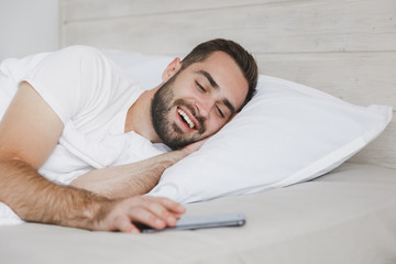 Calm young handsome bearded man lying in bed with white sheet pillow blanket in bedroom at home. Smiling beauty male spending time in room. Rest relax good mood lifestyle concept. Mock up copy space.