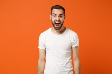 Angry crazy young man in casual white t-shirt posing isolated on bright orange wall background, studio portrait. People sincere emotions lifestyle concept. Mock up copy space. Screaming, swearing.