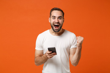 Shocked young man in casual white t-shirt posing isolated on orange background in studio. People lifestyle concept. Mock up copy space. Using mobile phone, typing sms message, doing winner gesture.