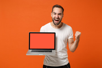 Excited young man in casual white t-shirt posing isolated on orange background. People lifestyle concept. Mock up copy space. Holding laptop pc computer with blank empty screen, doing winner gesture.