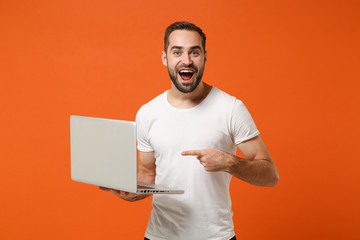 Excited young man in casual white t-shirt posing isolated on orange background studio portrait. People sincere emotions lifestyle concept. Mock up copy space. Point index finger on laptop pc computer.