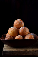 Donut balls in a clay bowl sprinkled with sugar powder on the edge of the wooden table against black background