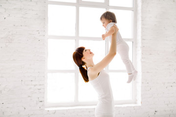 happy charming sporty mother in white wear holding cute little baby girl up overhead, while standing against big window in light hall gym