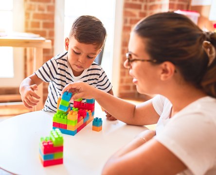 Beautiful teacher and toddler boy playing with construction blocks bulding tower at kindergarten