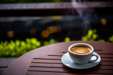 Hot black coffee with white smoke in a white cup, placed on a white saucer on a brown table in a coffee shop.