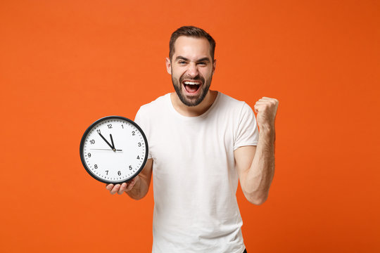 Happy young man in casual white t-shirt posing isolated on bright orange background studio portrait. People sincere emotions lifestyle concept. Mock up copy space. Holding clock, doing winner gesture.