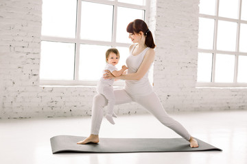 Obraz premium Health and fit concept. Young beautiful Caucasian woman doing fitness exercise in the big light gym with her little kid girl together. FItness materinity and motherhood