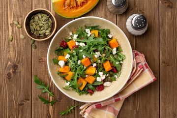 Autumn salad with baked pumpkin, arugula, seeds, dried cranberries and feta cheese in bowl on rustic wooden background. Top view.