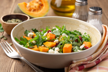 Autumn salad with baked pumpkin, arugula, seeds, dried cranberries and feta cheese in bowl on rustic wooden background. Selective focus.