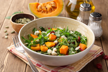 Autumn salad with baked pumpkin, arugula, seeds, dried cranberries and feta cheese in bowl on rustic wooden background. Selective focus.