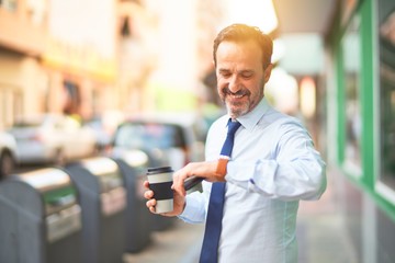 Middle age handsome businessman standing on the street drinking coffee looking at the watch smiling