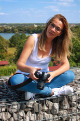 Fototapeta na wymiar Long-haired blonde sexy woman with a SLR camera. Girl in white t-shirt and blue jeans, sitting cross-legged on a low wall in the sun.